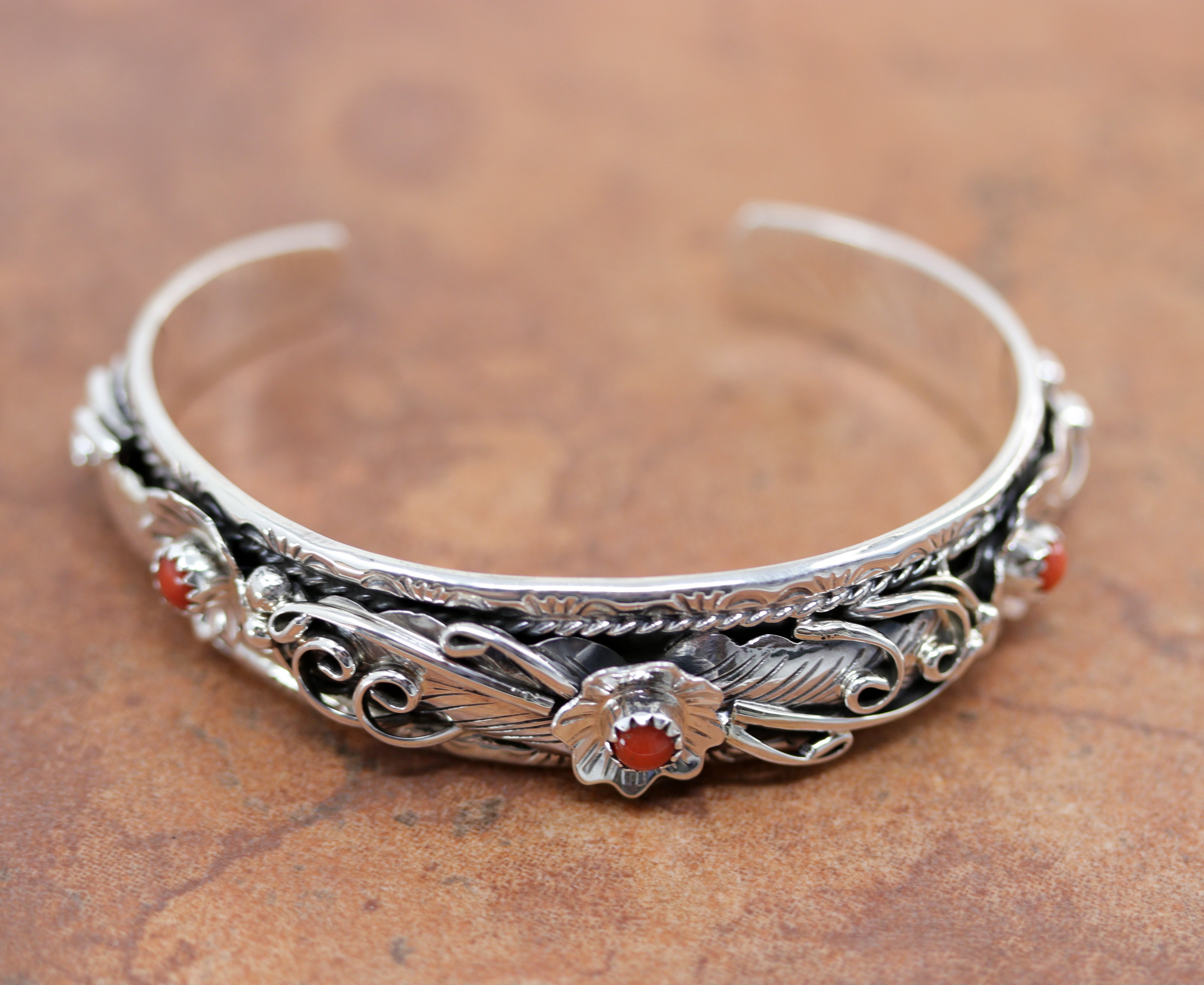 Silver cuff bracelet with floral and feather patterns. It has 3 circular red gemstones in the center of the flowers